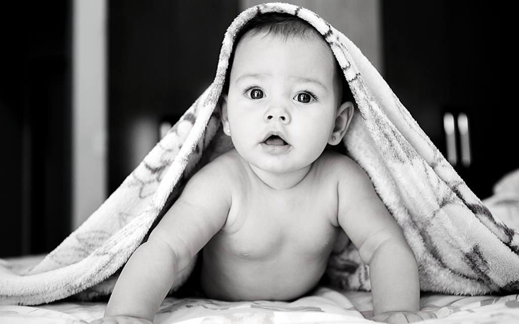 A baby girl with a blanket on top of her head looks straight into the camera. 