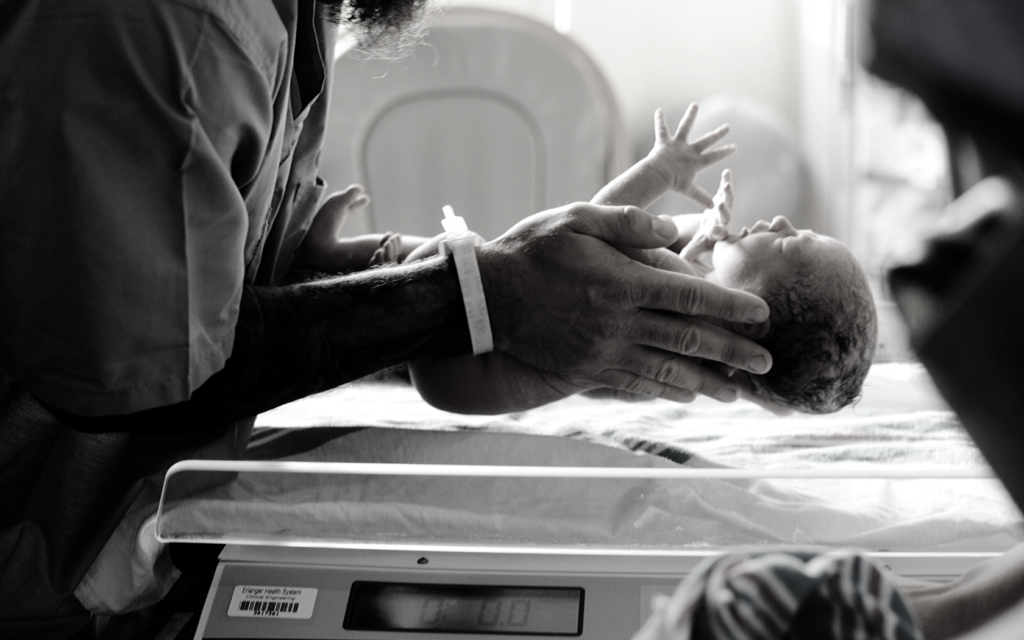A man wearing scrubs holds a newborn baby above a scale. 