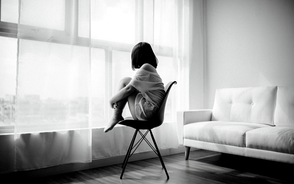A woman stares out the window as she sits in a chair with her knees cradles against her chest. 