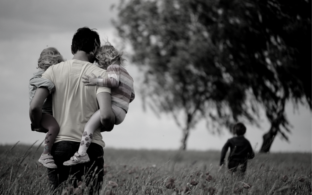 A father carries both of his children through a field of flowers. 