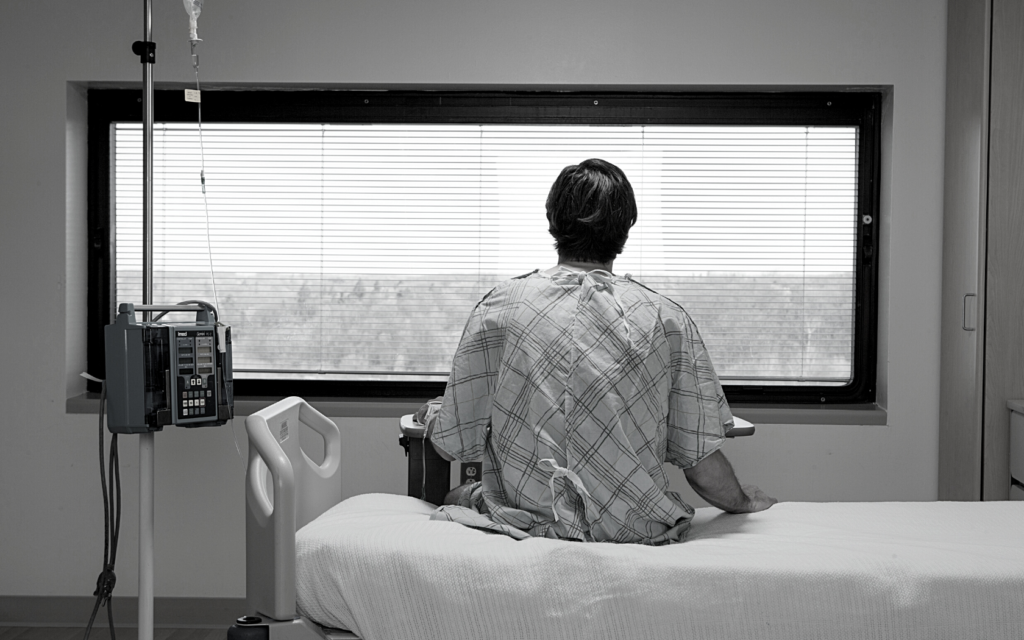 A man in a hospital gown sitting on a hospital bed stares out the window.