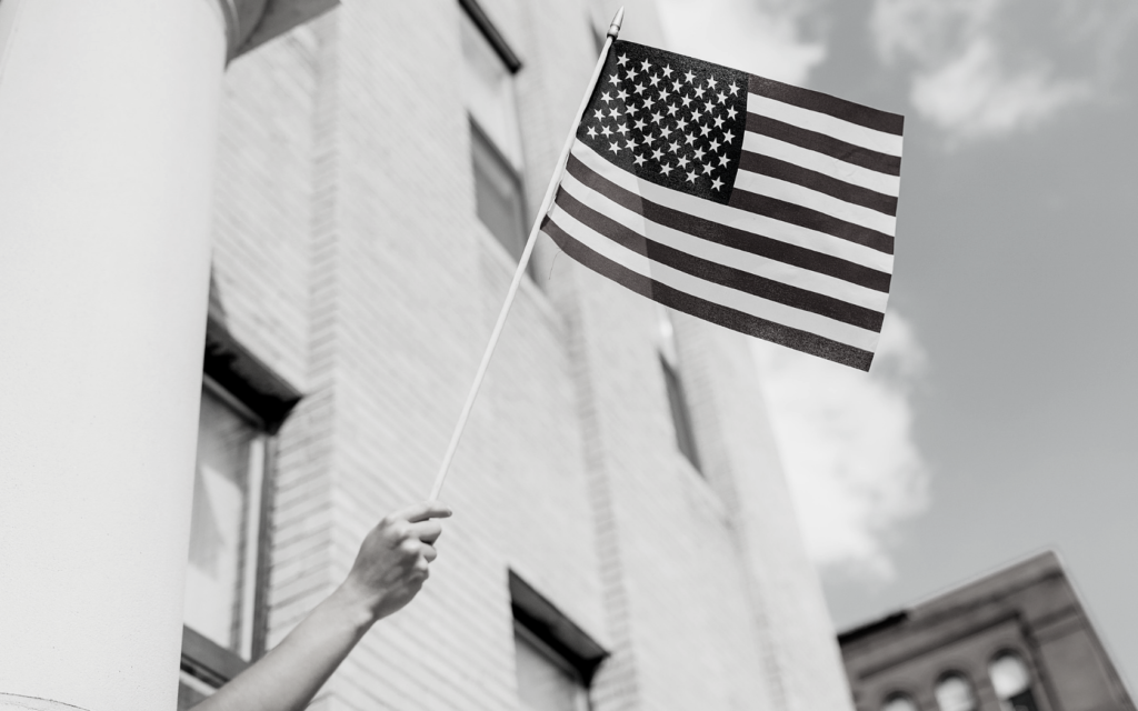 A woman inside a tall building holds an American flag outside window. 