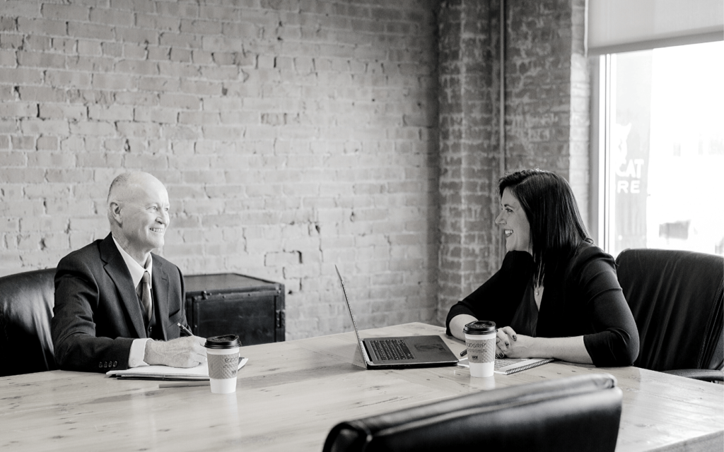 An older man and a younger woman sit at a desk with their laptops and have a professional conversation.